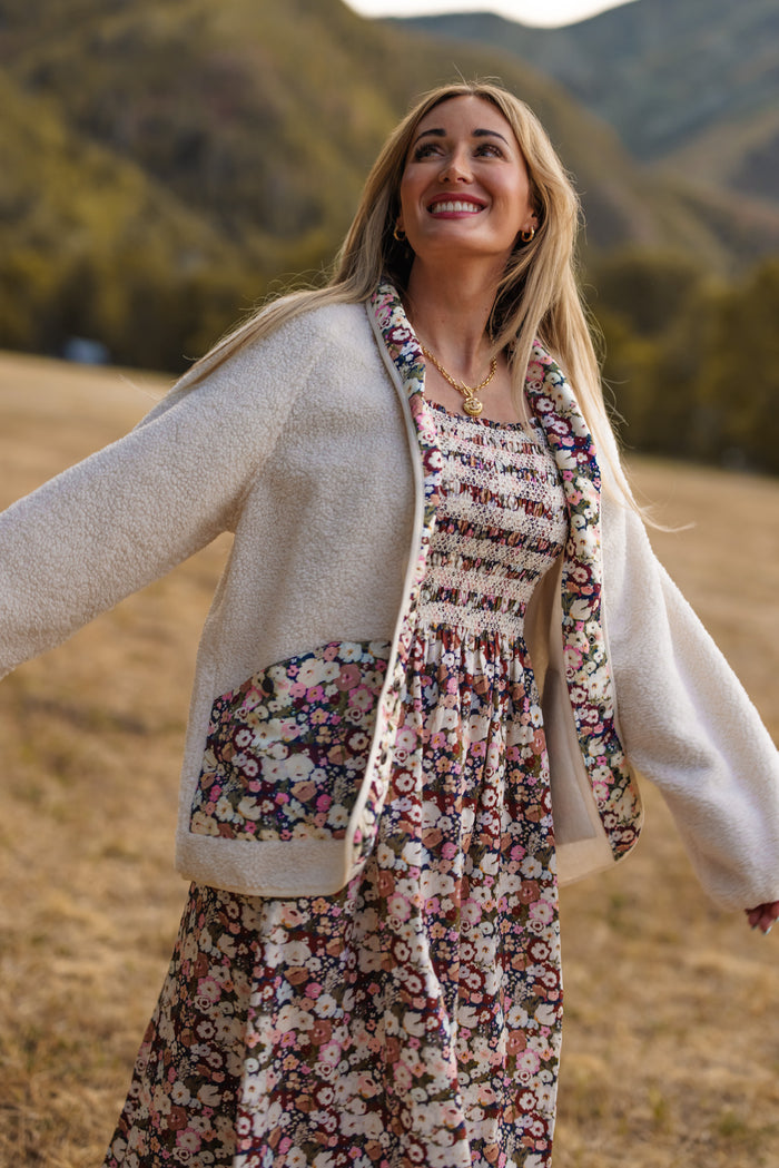 A woman smiles and spreads her arms while standing in a grassy field, wearing a floral dress and the Britta Jacket in Ivory & Midnight Garden by Ivy City Co, featuring delicate floral trim—an ideal addition to any fall wardrobe. Mountains and trees are softly blurred in the background.