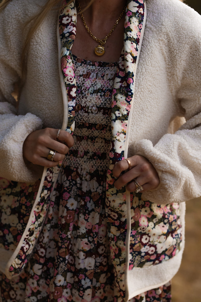 Wearing the Ivy City Co Britta Jacket in Ivory & Midnight Garden, styled over a floral dress, this person holds the sherpa-lined jacket open to reveal gold jewelry and rings on both hands—an effortlessly stylish look that’s perfect for any fall wardrobe.
