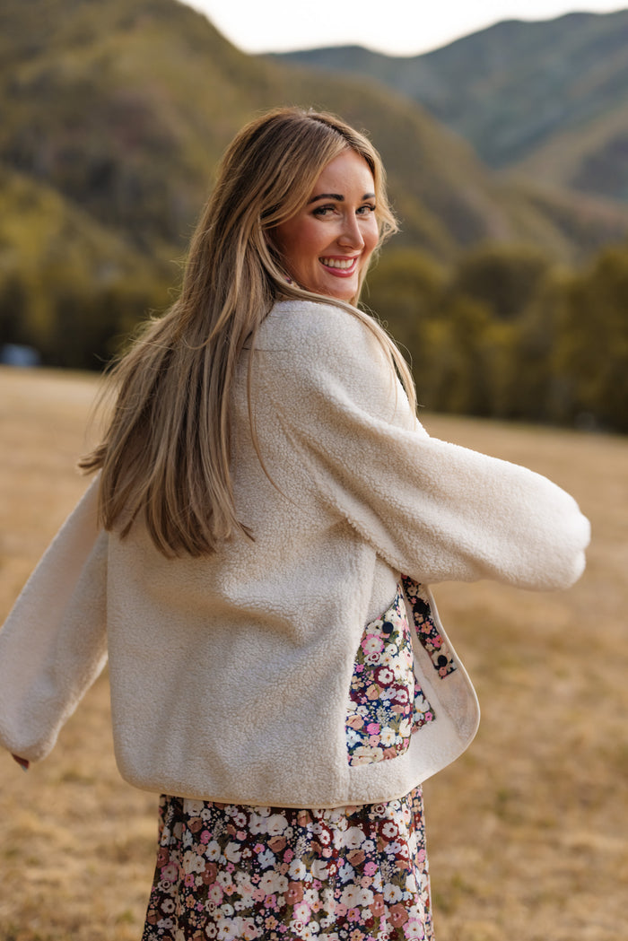 A smiling woman with long blonde hair wears the Britta Jacket in Ivory & Midnight Garden by Ivy City Co over a floral dress, looking back over her shoulder in a grassy field with rolling hills behind her—an ideal scene to inspire your fall wardrobe.