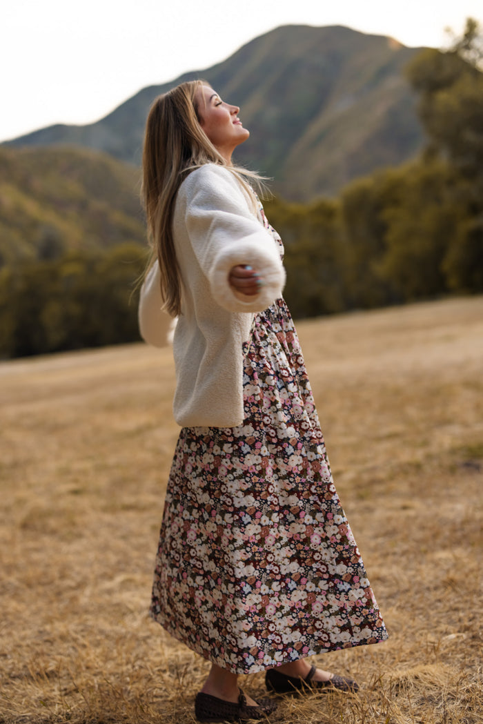 A woman wearing the Britta Jacket in Ivory by Ivy City Co and a Midnight Garden floral dress stands outdoors on dry grass with arms outstretched, smiling and looking up, framed by tree-covered mountains in the background—a perfect look for your fall wardrobe.