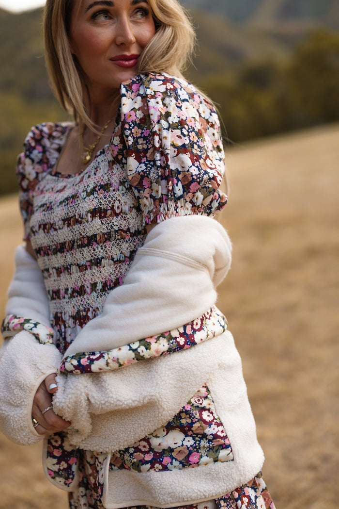 A woman stands outdoors wearing Ivy City Co’s Britta Jacket in Ivory, layered over a floral Midnight Garden dress. She gazes to the side, framed by blurred trees and hills—a perfect inspiration for your fall wardrobe.