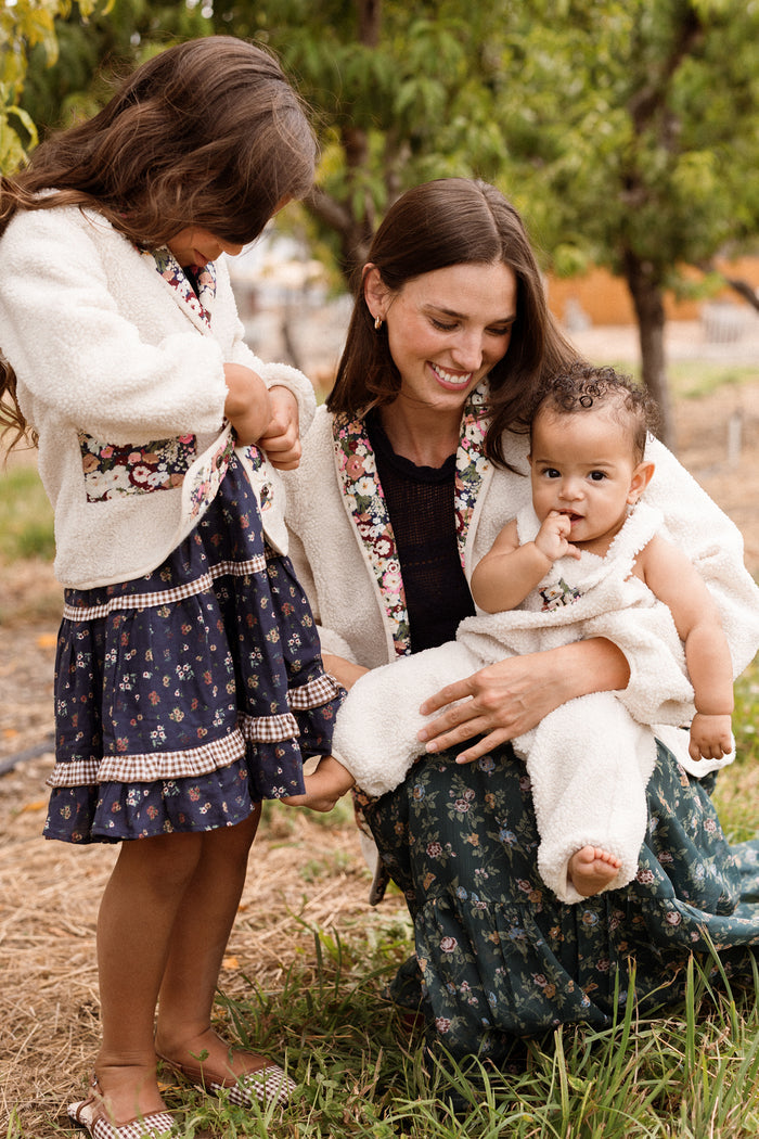 A woman smiles while holding a baby and sitting next to a young girl. All three wear floral-patterned outfits and are outdoors, surrounded by grass and trees. She completes her fall look with the Britta Jacket in Ivory & Midnight Garden by Ivy City Co, adding a cozy touch to her wardrobe.