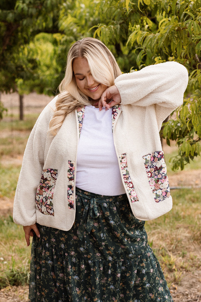 A smiling woman stands among green trees, tilting her head and touching her hair, wearing Ivy City Co’s Britta Jacket in Ivory & Midnight Garden—a cozy sherpa jacket with floral pockets that’s perfect for your fall wardrobe—paired with a white top and a dark green floral skirt.