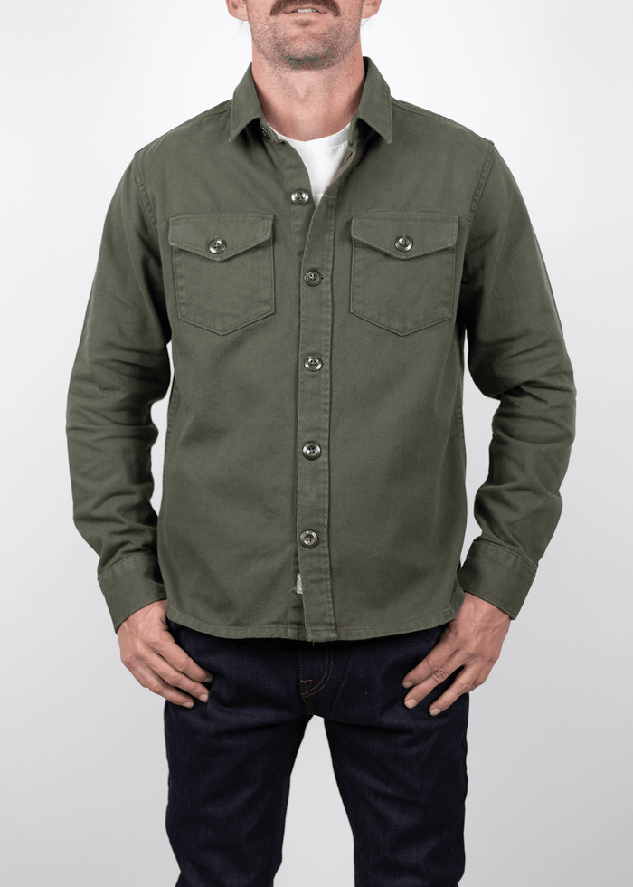 Man in olive green button-up shirt and dark jeans stands against a plain white background, hands resting casually by his sides.