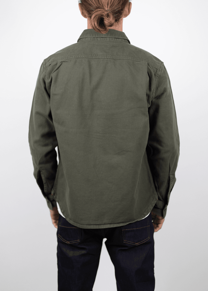 Man with a bun in a green jacket and dark jeans, facing away against a plain white background.