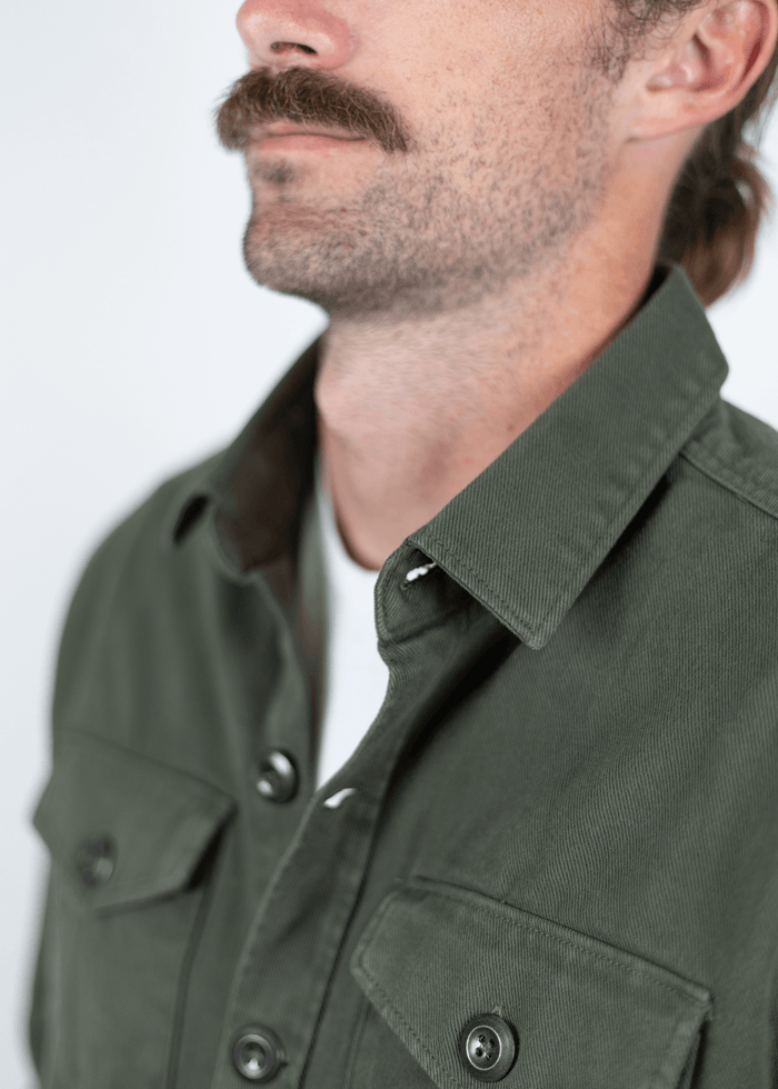 Man with mustache in green buttoned shirt, white undershirt, close-up on torso and face, neutral background.