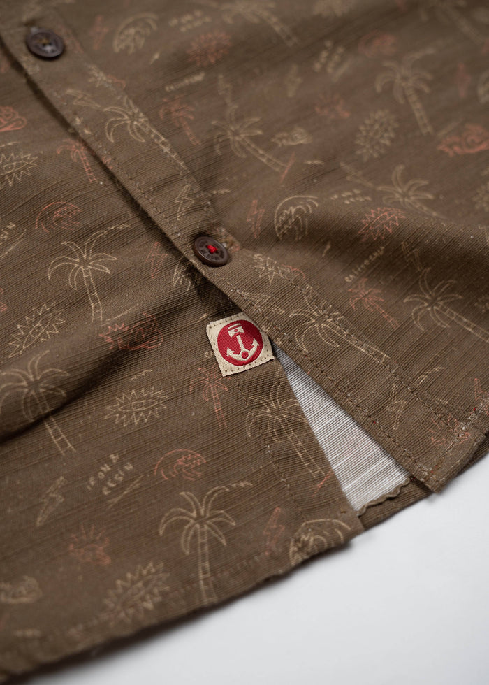 Brown shirt with palm tree pattern, close-up of buttons and red anchor tag detail, showcasing casual tropical style.