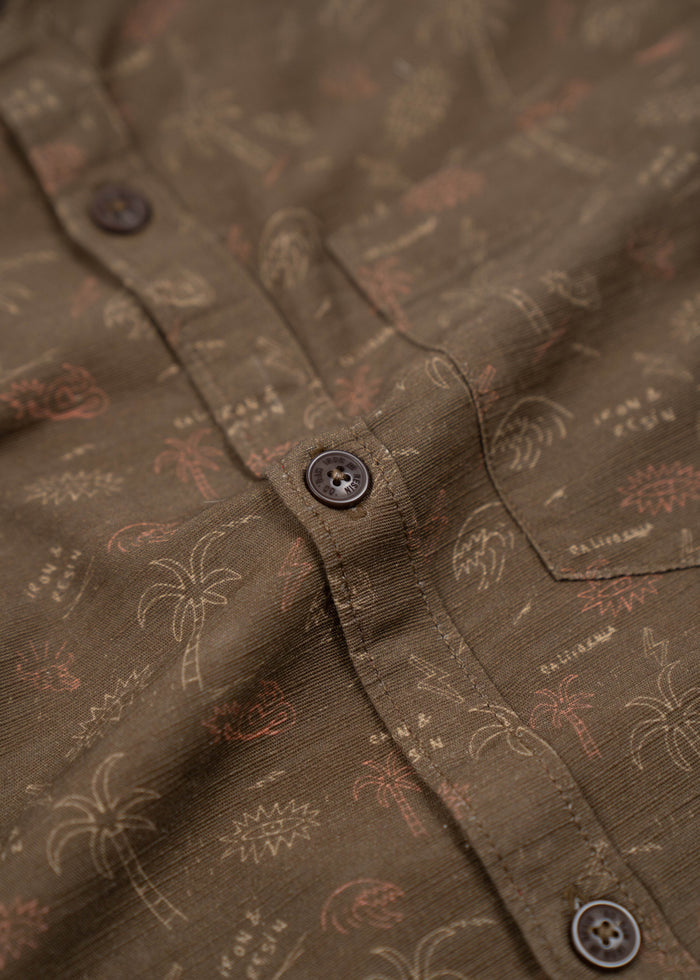 Close-up of a brown shirt with palm tree and text patterns, featuring dark buttons and a casual, tropical design.