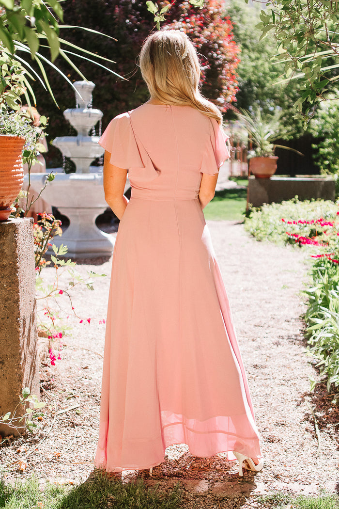 A woman with long blonde hair, wearing the Ivy City Co Callie Dress in Blush Chiffon, stands on a garden path facing away from the camera, with lush greenery and a white fountain in the background.