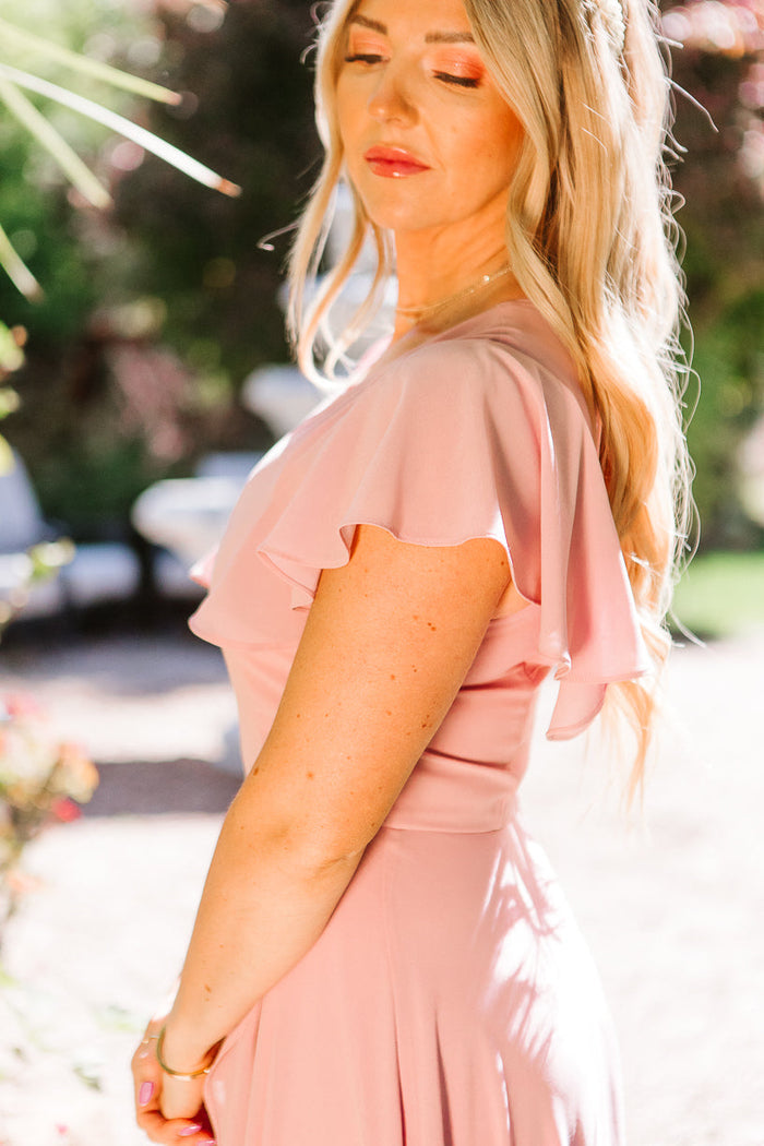 A woman with long blonde hair stands outdoors with her eyes closed, softly lit by sunlight and surrounded by greenery, wearing the Ivy City Co Callie Dress in Blush Chiffon.