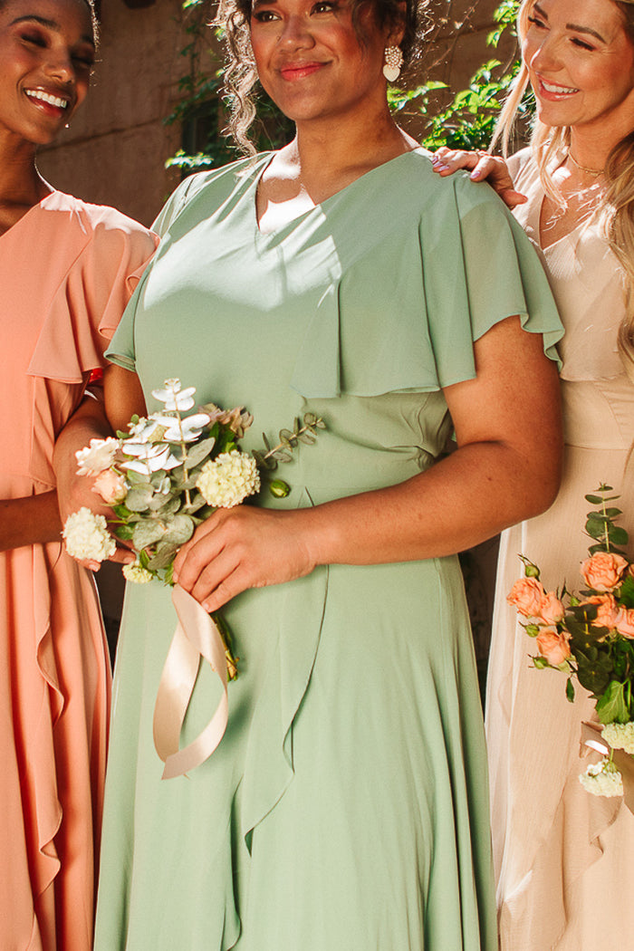 Three women in pastel wedding attire smile together, holding bouquets. The center woman wears the Ivy City Co Callie Dress in Sage Chiffon, while the others wear peach and beige maxi dresses. Sunlight filters through greenery behind them.