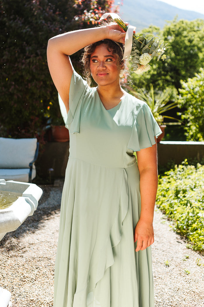 A woman with curly hair stands by a stone fountain in sunlight, holding a bouquet overhead. She wears the Ivy City Co Callie Dress in Sage Chiffon, surrounded by greenery.