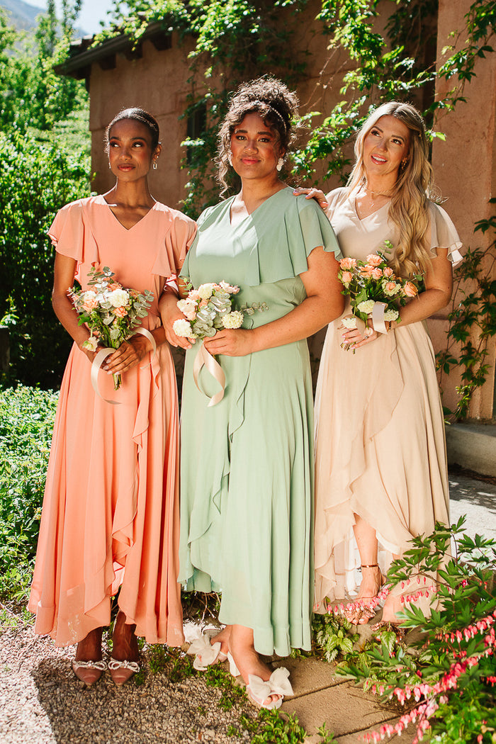 Three women wear Ivy City Co's Callie Dress in Sage Chiffon, standing outdoors with bouquets, smiling together amid greenery and sunlight, with a rustic building behind them.