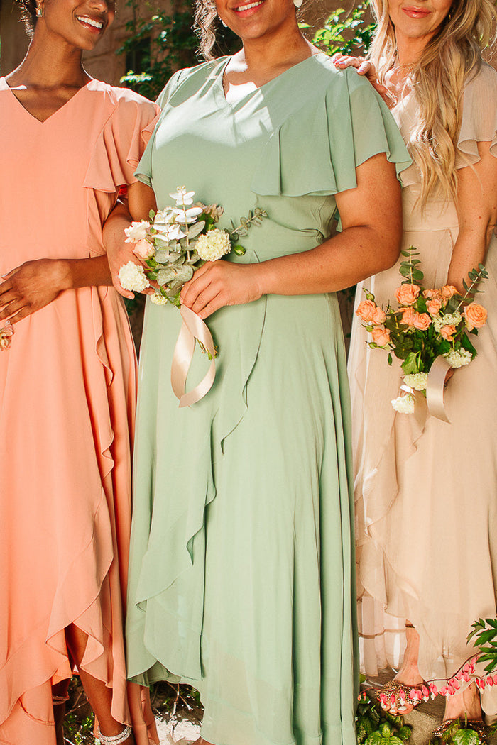 Three women in pastel dresses holding flower bouquets stand together outdoors. Only their bodies and part of their faces are visible; the central woman wears the Callie Dress in Sage Chiffon by Ivy City Co, while the others wear peach and beige maxi bridesmaid dresses.