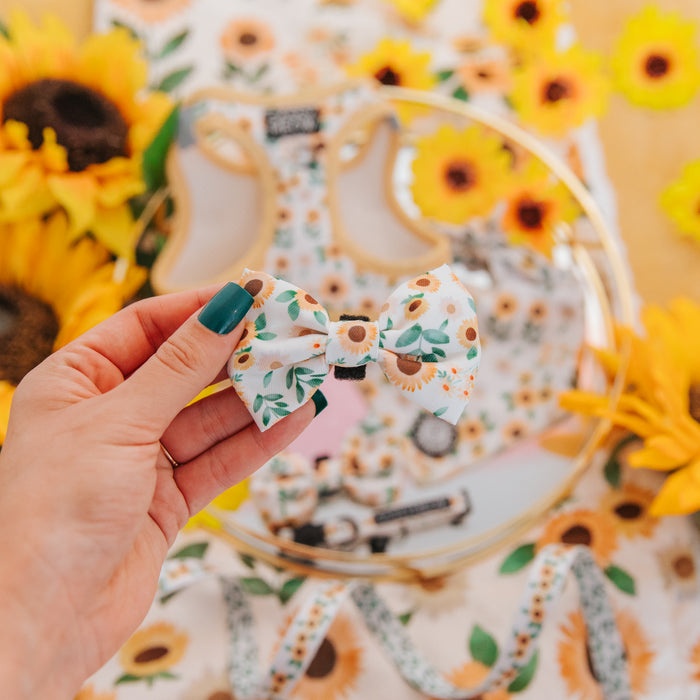 A hand with green nail polish holds the Sassy Woof Cat Bowtie - Sunflower Dreams. In the background, matching sunflower-themed cat accessories and a collar are displayed among fresh sunflowers, creating a vibrant scene.