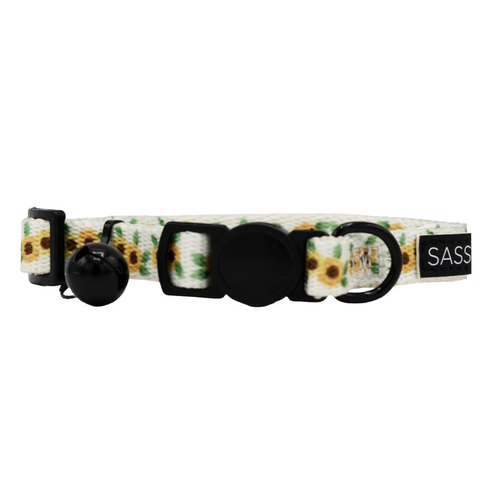 The Sassy Woof Cat Collar - Sunflower Dreams features a white sunflower design, black plastic buckle, adjustment slider, bell, tag holder, and a SASS label—perfect as a stylish and adjustable accessory for your cat.
