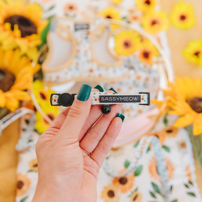 A hand with green nail polish holds the Sassy Woof 'Cat Collar - Sunflower Dreams,' featuring a floral pattern. Sunflowers and coordinating cat accessories appear blurred in the background.