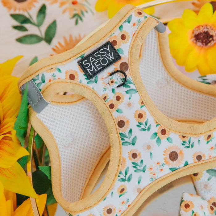 A close-up of the Sassy Woof Cat Step-In Harness - Sunflower Dreams, featuring an AirTag pocket, yellow trim, and a sunflower with green leaf design, is surrounded by bright sunflowers for added charm.