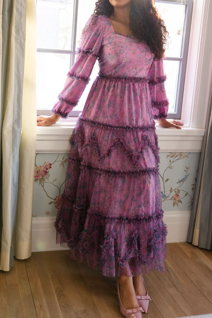 A woman in the Ivy City Co Catherine Dress in Climbing Vine Bloom stands by a window, light shining behind her pink heels. Floral wallpaper and wood floors complement her elegant spring look.