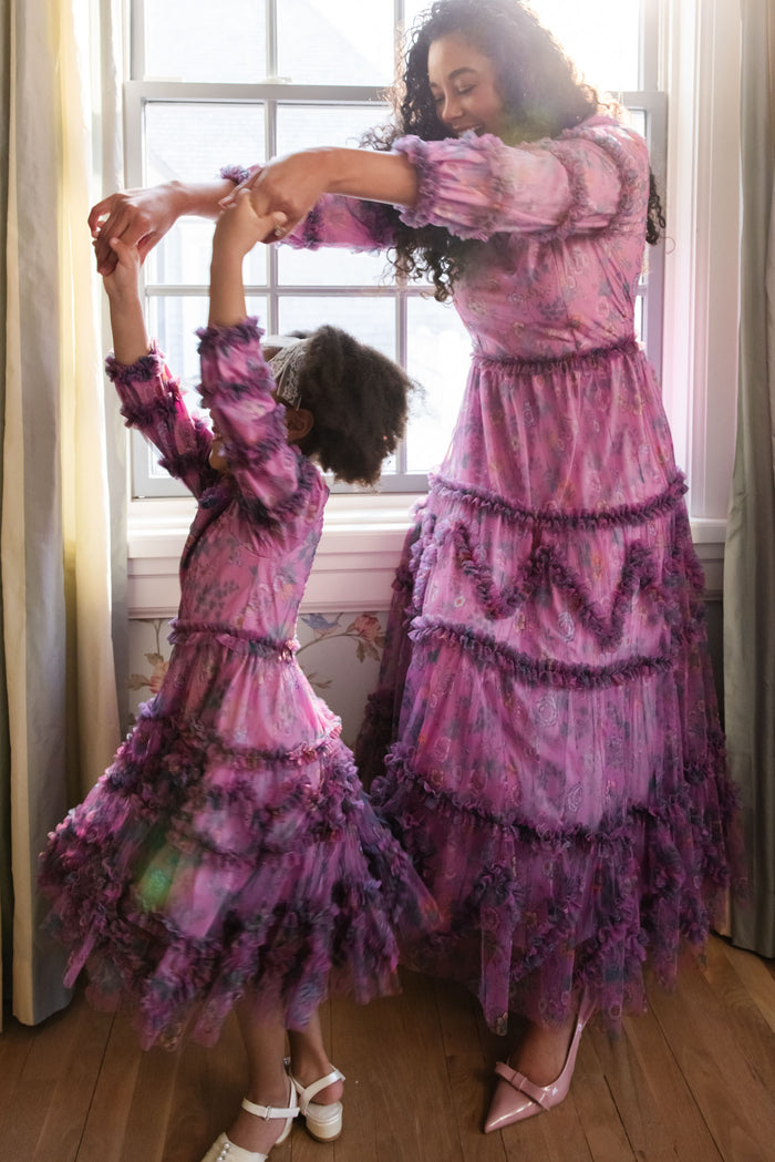 A woman and a young girl, both in matching Ivy City Co's Catherine Dress in Climbing Vine Bloom, dance hand-in-hand by a sunlit window, sharing smiles and enjoying the moment in their elegant spring outfits.