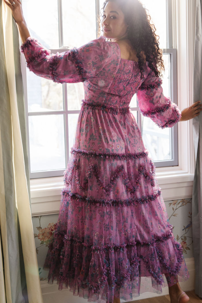 A woman with curly hair stands by a window, looking over her shoulder in the sheer purple Catherine Dress in Climbing Vine Bloom by Ivy City Co, its puffed sleeves and layered skirt glowing as sunlight streams through behind her.