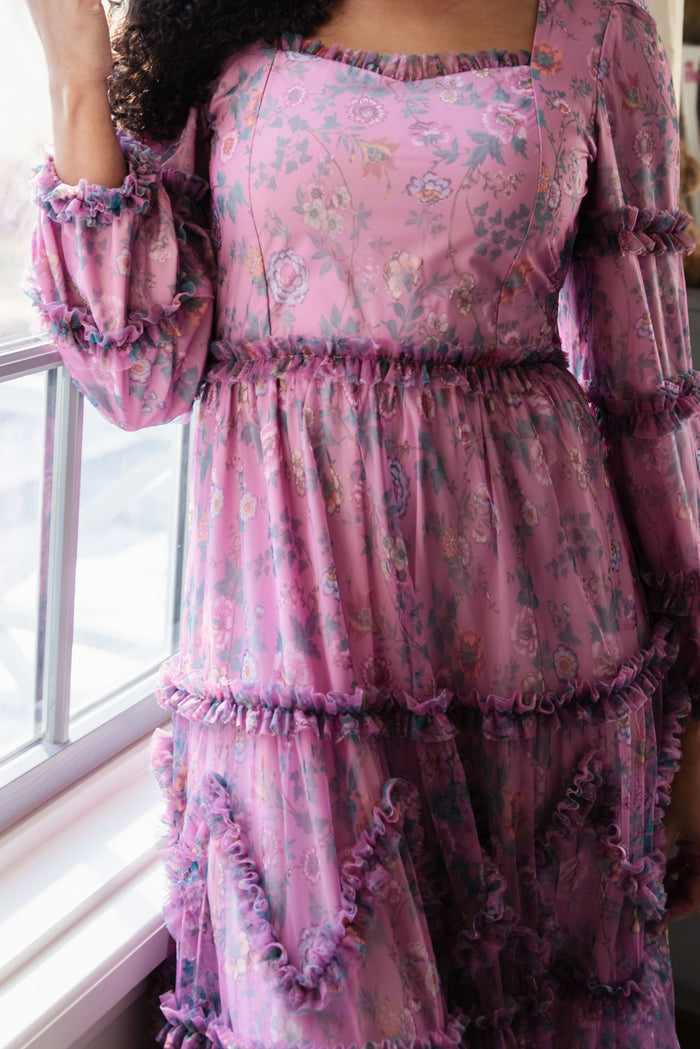A person stands by a window wearing the Ivy City Co Catherine Dress in Climbing Vine Bloom, a pink ruffled long-sleeve spring dress with sheer layers and purple trim. Only the upper body is visible, bathed in soft window light.