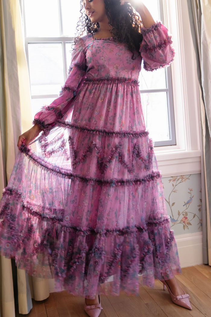 A woman indoors by a window wears the Ivy City Co Catherine Dress in Climbing Vine Bloom with pink heels, holding out one side to showcase its elegant, flowing look as sunlight streams in behind her.