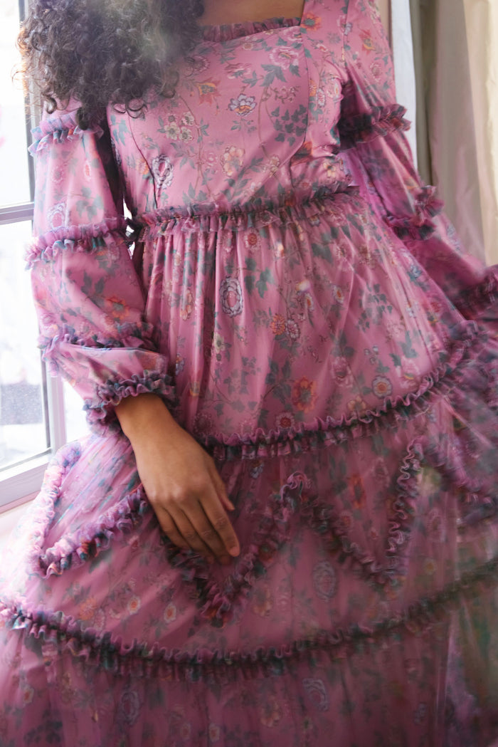 A person wearing the Ivy City Co Catherine Dress in Climbing Vine Bloom, featuring puffed sleeves and ruffles, stands near a window. The upper part of their face is not visible in the image.