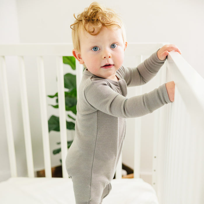 Child in a Dove color footless baby romper standing in a crib with a white background