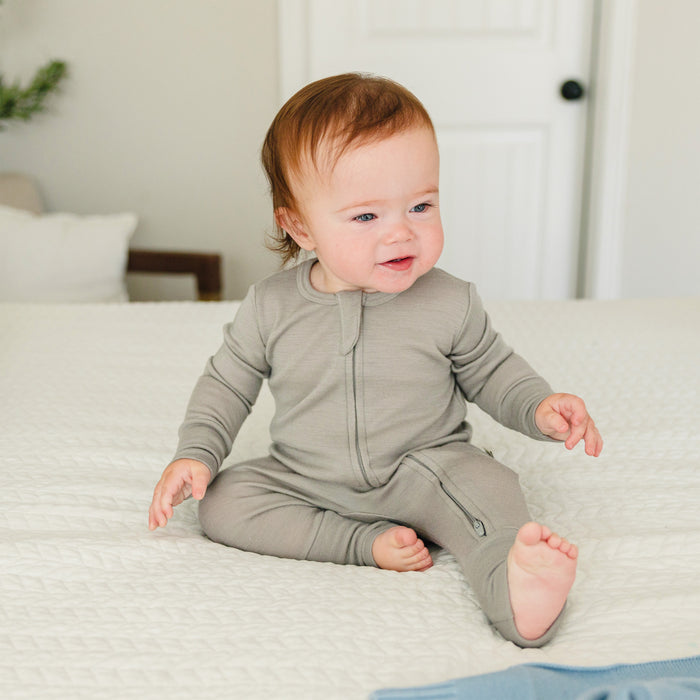 Baby in a dove-colored footless baby romper sitting on a bed in a home setting