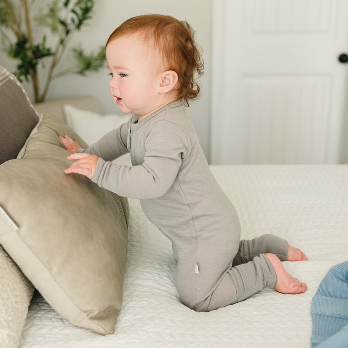 Baby in a Dove color footless baby romper sitting on a couch with pillows