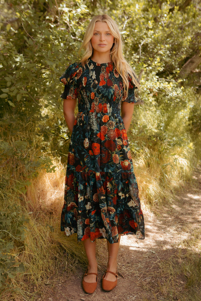 A woman with long blonde hair stands outdoors on a dirt path, wearing the Ivy City Co Cordelia Dress in Moody Floral—a dark floral chiffon midi dress with short ruffled sleeves—paired with brown flat shoes. Green foliage and sunlight surround her, making this look perfect for a maternity-friendly fall dress style.