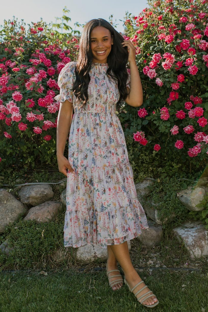 A woman in the Ivy City Co Cordelia Floral Dress with a smocked bodice smiles, standing on grass with one hand in her hair, surrounded by blooming pink roses, greenery, and rocks at her feet.