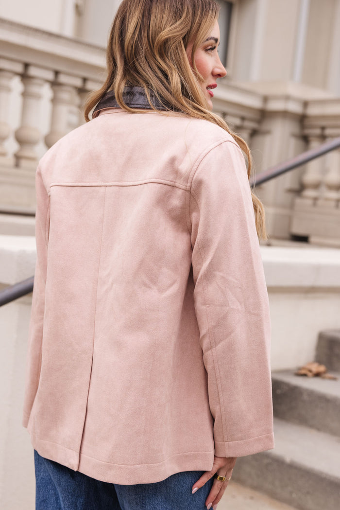 A woman with long, wavy hair stands outdoors facing away, wearing the Ivy City Co Daphne Coat in Light Pink and blue jeans near a staircase with a decorative railing and a neutral building in the background.