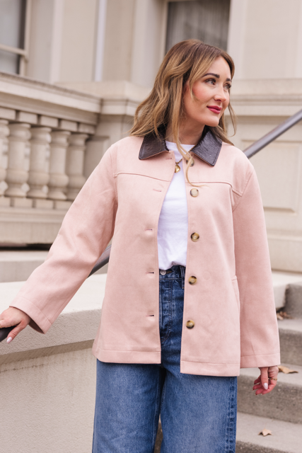 Wearing the Ivy City Co Daphne Coat in Light Pink over a white shirt and blue jeans, a woman stands near stairs outside a building, looking to the side.