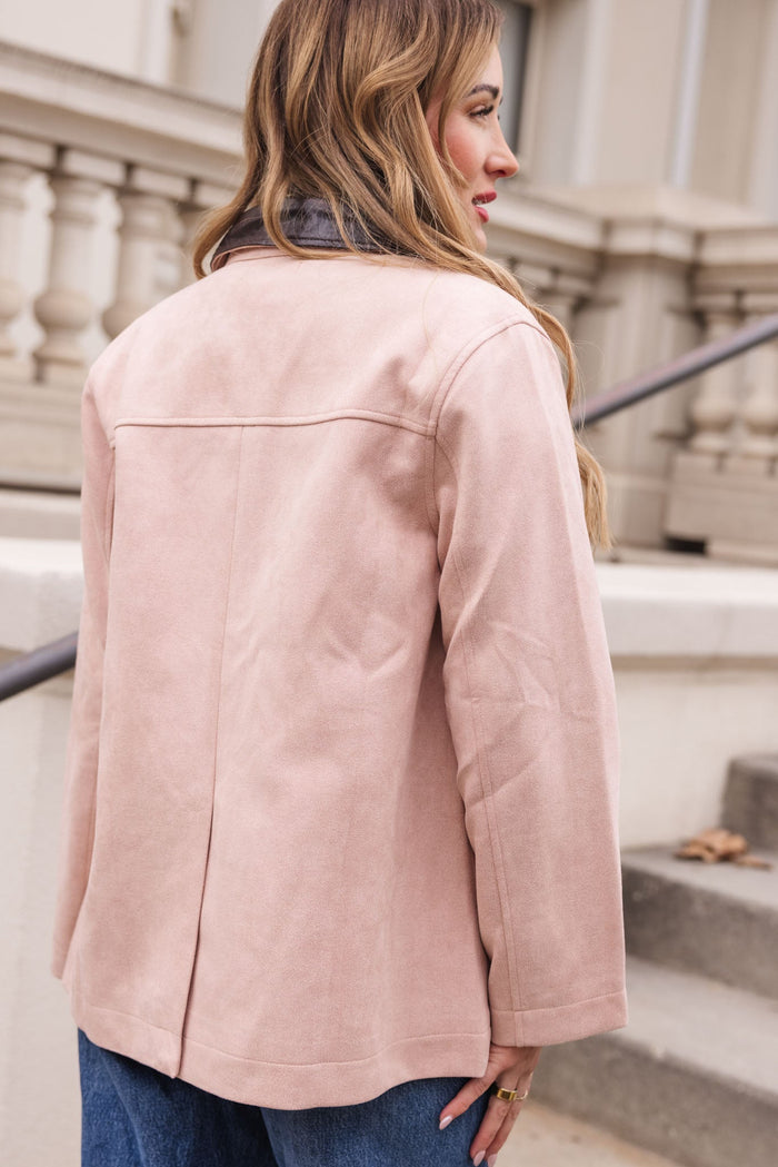 A woman stands outdoors facing away, wearing the Ivy City Co Daphne Coat in Light Pink paired with blue jeans. Her long, wavy hair cascades over her shoulder, and a stone railing and steps are visible in the background.