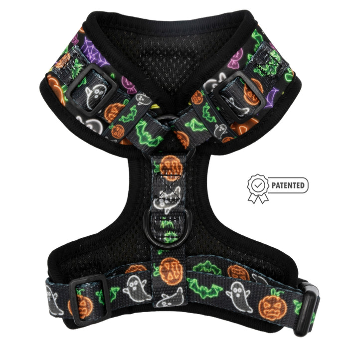 The Sassy Woof Dog Adjustable Harness - Neon Frights is a black mesh harness with neon prints of ghosts, pumpkins, bats, and spider webs. It features sturdy straps, a D-ring for leash attachment, and a patented icon beside it.