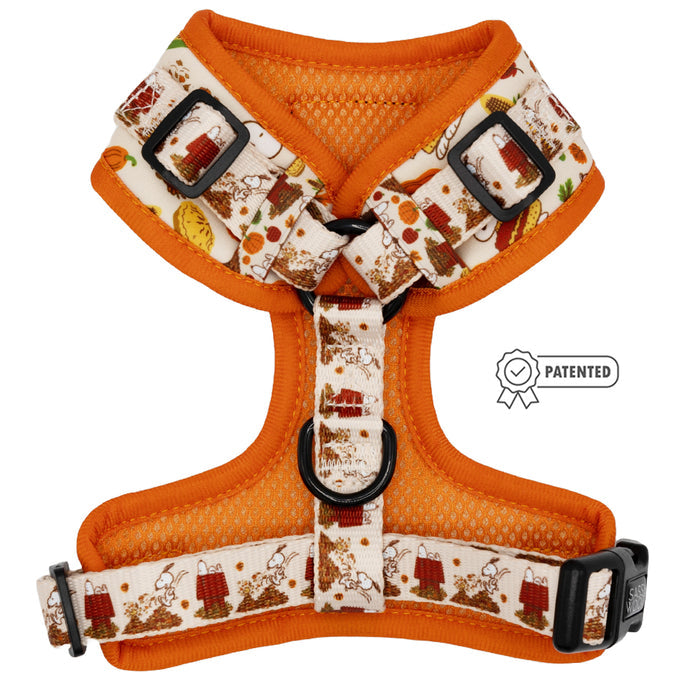 The Sassy Woof Dog Adjustable Harness - Peanuts™ Harvest features an orange body with white straps, brown and red cartoon Snoopy designs, black adjustable buckles, a center D-ring, and a patented badge on the right side.