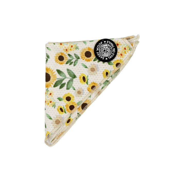 The Dog Bandana - Sunflower Dreams by Sassy Woof features a folded yellow and white design with sunflower and greenery patterns. Displayed on a white background, it includes a black "Stay Wild Woof Sassy" tag and pairs perfectly with outdoor dog accessories.