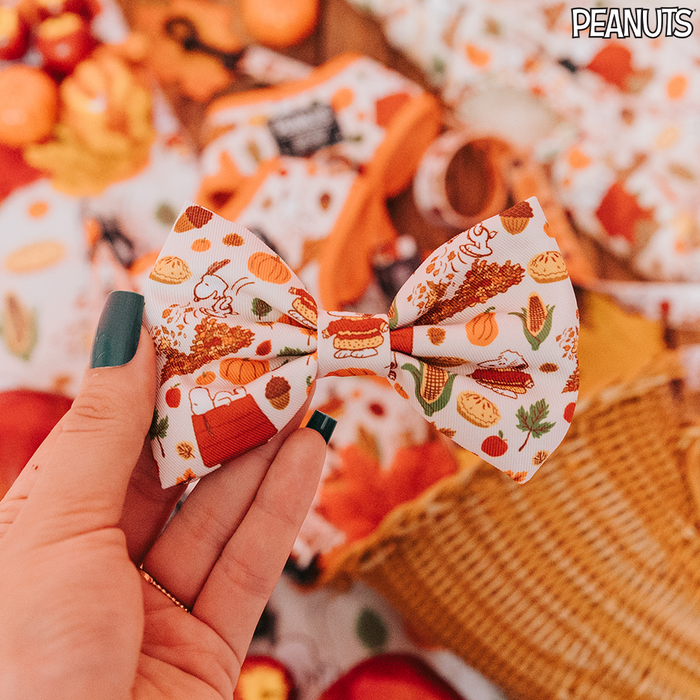 A hand with teal nail polish holds the Sassy Woof Dog Bowtie - Peanuts™ Harvest, decorated with Snoopy, pumpkins, and fall treats. The backdrop features warm seasonal colors and matching festive items.