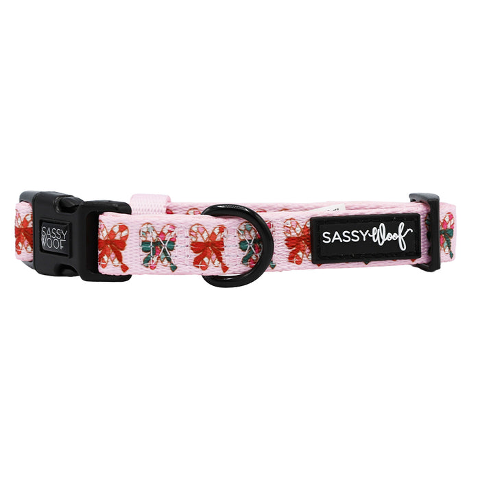 The Dog Collar - Candy Cane Coquette by Sassy Woof features a pink festive design with red and green bows, a black buckle, metal ring, brand logo patch, and adjustable fit—ideal for holiday accessories.