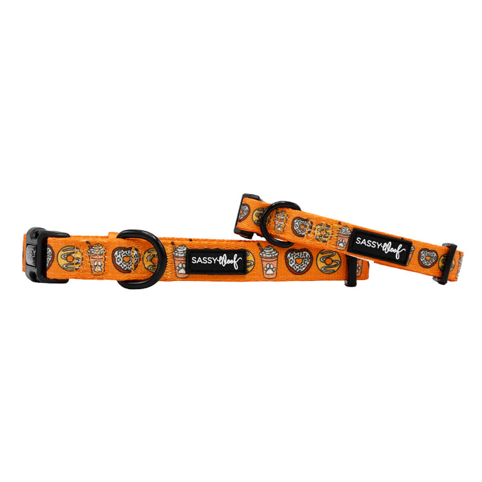 Two Sassy Woof "Pupkin Donuts" dog collars in orange with black buckles, metal rings, and Halloween cartoon designs are displayed on a white background. Both collars feature the Sassy Woof logo patch.