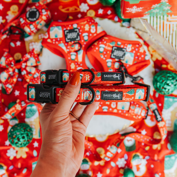 A hand with orange-painted nails holds two red Sassy Woof Dog Collar - Sleigh Baby Sleigh, featuring festive holiday dog accessories and blurred matching decorations in the background.