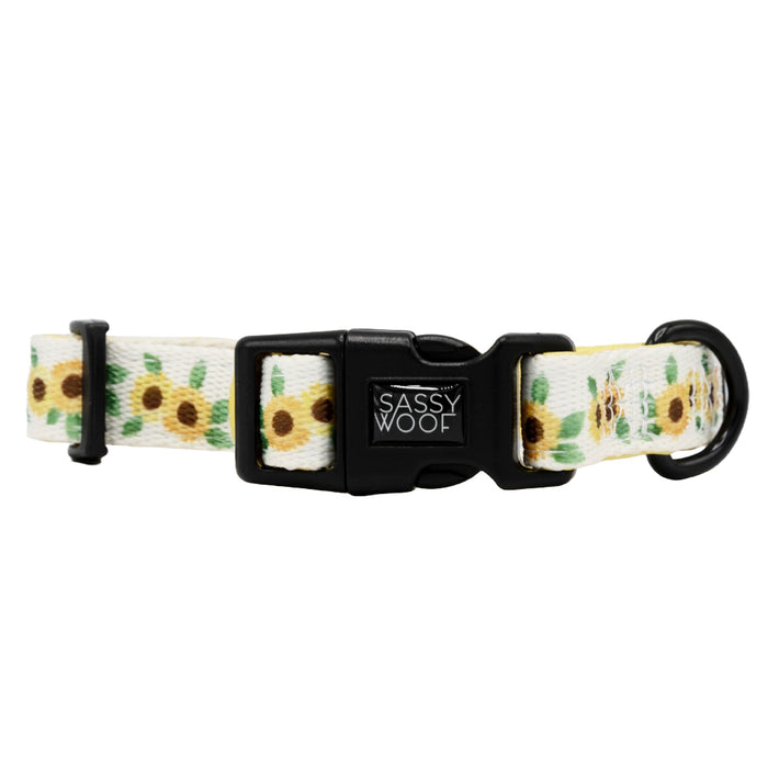 The Dog Collar - Sunflower Dreams by Sassy Woof is a white adjustable collar with a vibrant sunflower pattern, black plastic buckle with brand logo, and a sturdy black metal D-ring for leash attachment.