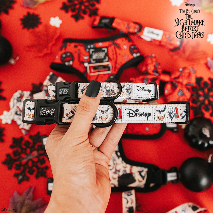 A hand holds two Sassy Woof Dog Collars – The Nightmare Before Christmas™ (Beige), featuring cartoon characters, set against a red background with themed pet accessories and black paper cutouts.