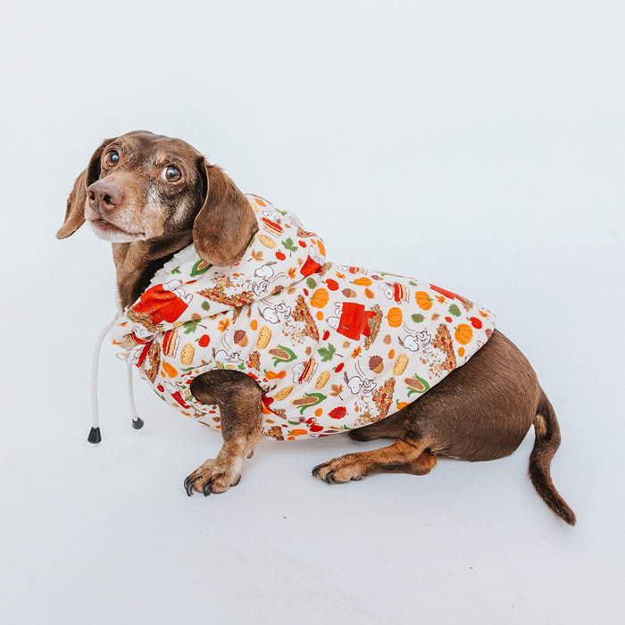A brown dachshund wears the Sassy Woof Dog Reversible Sherpa Jacket - Peanuts™ Harvest, a white hooded jacket with colorful food and vegetable illustrations, and sits on a white background looking back over its shoulder.