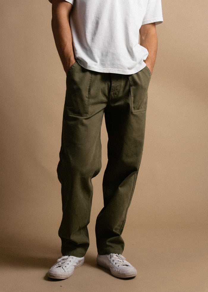 Person in a white shirt and olive green pants stands casually with hands in pockets, wearing white sneakers on a beige background.