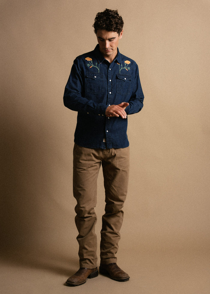 Man in embroidered denim shirt and brown pants stands against a beige background, looking down and adjusting his sleeve.