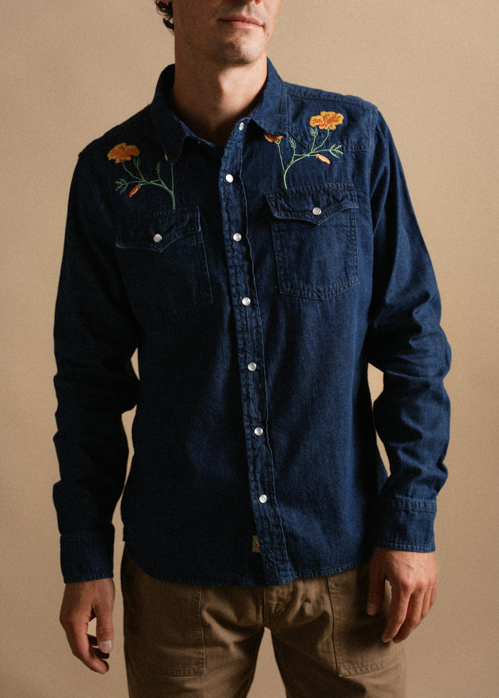 Man in a blue denim shirt with floral embroidery stands against a beige background, hands relaxed by his sides.