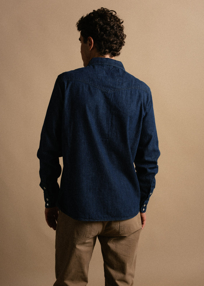 Man in a blue denim shirt and khaki pants stands against a beige background, facing away. Curly hair, casual style.